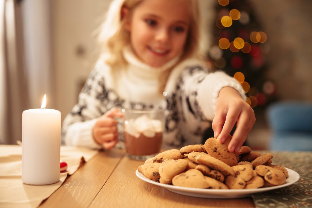 Little girl taking delicious cookie from plate while sitting at festive table, selective focus on hand